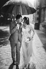 Wall Mural - Beautiful wedding couple embracing together under umbrella in sunny rainy street. Stylish bride and groom walking and kissing on background of old church in rain. Black and white photo