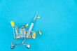 Shopping cart with medical ampoule, syringe and capsules on a blue background, copy space, top view. Concept of purchasing medicines, increasing drug prices