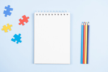 Wall Mural - Blank open spiral paper notebook with blue, yellow, red pieces of puzzle and colored pencils on light blue background. World autism awareness day concept. Top view, copy space