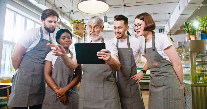 Portrait of busy senior Caucasian male restaurant owner in apron tapping on tablet looking at screen discussing with mixed-race young male and female employees working plan. Business concept