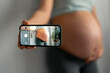 © Dorde - Unrecognizable pregnant woman with stretch marks capturing her baby bump, taking photos with the smartphone, wearing comfortable cloths, a blue t-shirt and gray leggings, holding her hand on stomach.