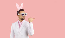 Young Handsome Man With Easter Rabbit Ears Wearing White Suit, Pink Shirt And Sunglasses Standing Over Isolated Pink Background Smiling With Happy Face Looking And Pointing To The Side With Thumb Up. 