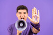 © Egoitz - Young male protestor shouting on megaphone and showing a ribbon drawn on hand isolated on purple background. Feminism, equality and gender violence concept.
