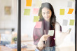 © David - Business female employee with many conflicting priorities arranging sticky notes commenting and brainstorming on work priorities colleague in a modern office.