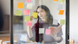 © David - Business female employee with many conflicting priorities arranging sticky notes commenting and brainstorming on work priorities colleague in a modern office.