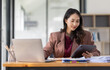© David - Freelancer Happy business Asian woman in knitwear taking notes at laptop sitting at desk office, finance concept.