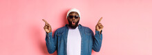 Amazed Cool Black Guy Showing Two Variants, Pointing Fingers Sideways And Staring At Camera Impressed, Wearing Sunglasses, Standing Over Pink Background