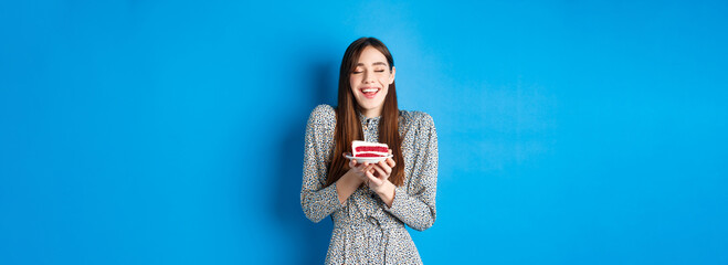 Dreamy birthday girl making wish with closed eyes and happy smile, blowing cake candle, standing on blue background