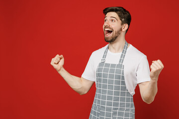 Wall Mural - Side view young male housewife housekeeper chef cook baker man wear grey apron do winner gesture celebrate clenching fists say yes isolated on plain red color background studio Cooking food concept