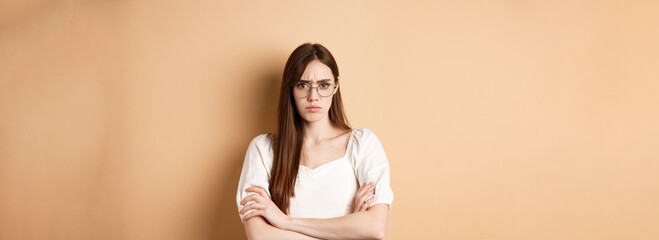 Angry and offended girl in glasses frowning, cross arms on chest and sulking, standing defensive on beige background