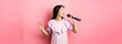 Beautiful asian girl perform song, singing in microphone and smiling romantic, standing in dress against pink background