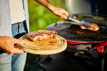 Wall Mural - Man put down on a wooden board ready to eat grilled steak meat, male roasted steak meat on the gas grill on barbecue grill outdoor in the backyard, summer family picnic, food on the nature.