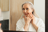 Fototapeta  - Senior woman with long grey hair touching her cheek with fingers in bathroom