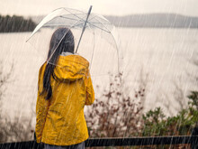 Teenager Girl In Yellow Jacket Holding Translucent Umbrella Looking At A Beautiful Lake In The Background In A Rain. Selective Focus. Outdoor Activity. Going Out.