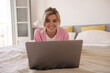Nice young caucasian girl lies on bed and works on laptop with wide smile at home. Blonde hair woman wears pink sweater and white t-shirt indoor. Concept positive mood while working.