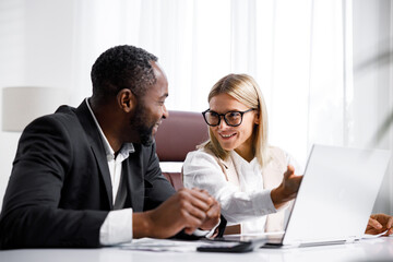 Two people analyze documents on a laptop while sitting at a table in the office. Business partners at work in the office are discussing projects. Multi-racial collaboration.