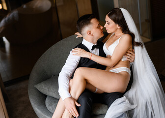 Above view of handsome groom in suit, holding bride on knees while sitting on comfortable chair in modern apartment. Sexual woman with curly hair,  wearing in lace lingerie and wedding veil