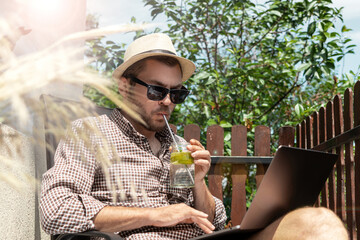 Handsome man with sunglasses and straw hat drinking fresh water with lime while sitting on terrace in summer using his laptop.