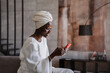 © Iona - Happy african young woman in white turban and white dress sits on cozy sofa at hotel, reads funny message from friend. Cheerful Brazilian girl surfing social media laughs. Grateful female chatting.