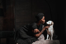 Young African Woman In Black Turban And Dress Sits On Cozy Chair With Little White Dog Strokes Puppy At Home. Owners And Pets. Domestic Animals. Pretty Brazilian Girl Taking Care Of Hound. Mockup