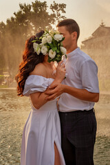 Groom gently hugs bride and they kiss covered with a bouquet of delicate white flowers. Newlyweds in white robes against the backdrop of splashes from the fountain and the setting sun.