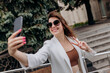 © bondvit - Smiling business woman in white suit and sunglasses making video call to friend using phone during break standing near modern office building