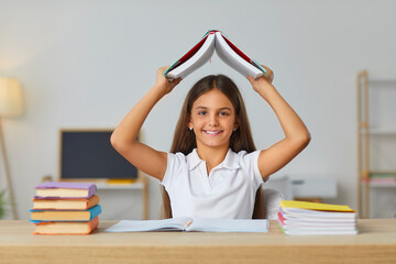 Portrait of happy school child with books. Beautiful student girl sitting at desk in classroom, looking at camera, smiling and holding book above her head like house roof top. Back to school concept