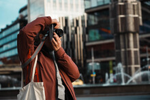 Blond Girl Takes Pictures With Analog Camera. Experienced Young Photographer Takes Souvenir Photos At Sunset In The City. Tourist In Red Jacket And Shoulder Bag Focuses On The Lens. Street Photography