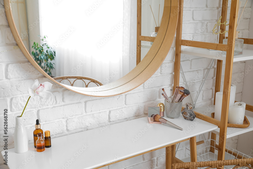 Table with makeup brushes, cosmetics and flower in vase near white brick wall