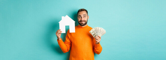 Real estate concept and mortgage concept. Handsome young man buying house, holding home maket and money, thinking and smiling, standing over light blue background