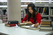 © PhotoFL - Chica universitaria de blusa roja escribiendo y estudiando en la biblioteca	mientras escucha música con sus audífonos