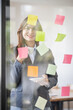 © David - Business Asian female employee with many conflicting priorities arranging sticky notes commenting and brainstorming on work priorities colleague in a modern office.