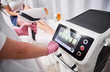 Close up of female beautician in sterile gloves using diode laser hair removal machine while woman sitting on daybed. Esthetician preparing equipment for epilation procedure.