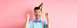 Holidays and celebration concept. Cheerful young man celebrating birthday party in cone hat and bow-tie, shouting yes with excitement and making fist pump gesture, pink background
