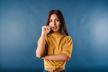 Portrait Of Brunette Woman Wearing Mustard Yellow T-shirt Isolated Over Blue Background Woman Biting Her Nails While Looking At Camera. Scared And Doesn't Know What To Do.