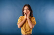 © platinumArt - Young beautiful woman wearing yellow top isolated over blue background looks at the camera with her mouth open and touches her face. Cannot believe it. Wow mouth.