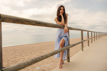 Wall Mural - Tender young caucasian woman with wavy hair relaxing on beach in cloudy weather. Brunette wears striped sun-dress looking at camera. Concept coastal weekend.