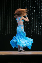 A Young Dancer With Long Hair In An Oriental Costume With A Multi-colored Shawl Poses And Dances A Belly Dance On A Concert Stage. Professional Stage Lighting.