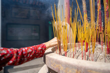 Close-up Of A Hand Holding Incense. The Background Is An Unspecified, Faded Temple