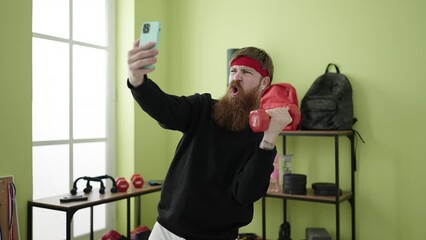 Wall Mural - Young redhead man using dumbbells training and making selfie by smartphone at sport center