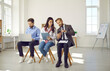 © Studio Romantic - Business people waiting for job interview, front view. Applicants sitting on chairs in queue preparing for interview, candidates holding resume using smartphones and laptop. Hiring and job search