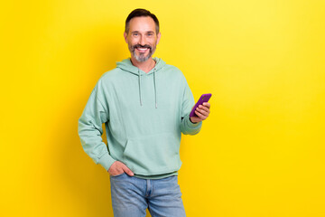 Canvas Print - Photo of satisfied technology gadget user middle age man wear khaki hoodie hold phone browsing new dating app isolated on yellow color background