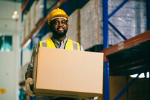 Worker In Warehouse Holding Cardboard Box While Looking At Camera And Smiling..