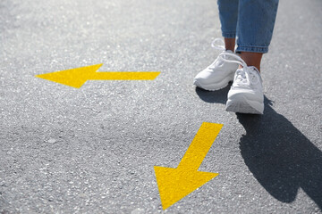 Wall Mural - Choice of way. Woman walking to drawn mark on road, closeup. yellow arrows pointing in different directions
