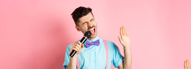 Party and festive events concept. Funny guy performs a song, singing karaoke in microphone with carefree face, standing in bow-tie and suspenders on pink background