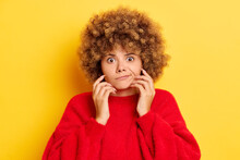 Portrait Shot Of Stunned Emotional Young Woman With Curly Hair Keeps Fingers On A Face And Looks Frightened, Wears Red Pullover, Poses Over Yellow Wall