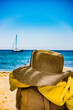 Un chapeau en paille est posé sur une chaise sur une plage  en été devant une mer bleu turquoise , on devine un voilier au loin