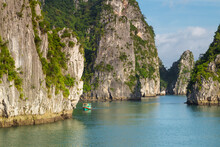 A Small Green Boat Dwarfed By Towering Limestone Karsts Coming Out Of The Water At Ha Long Bay In Vietnam