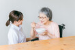 老人ホーム・家で食事する笑顔の高齢者女性と食事介助する管理栄養士・介護士・ヘルパーのアジア人女性