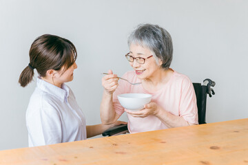 Wall Mural - 老人ホーム・家で食事する笑顔の高齢者女性と食事介助する管理栄養士・介護士・ヘルパーのアジア人女性